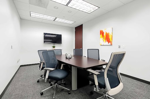 Small modern conference room with a dark wooden table, six mesh chairs, wall-mounted TV, abstract painting, and a doorway.