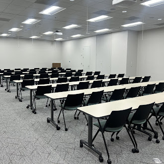 Empty classroom with rows of white desks and black wheeled chairs under bright ceiling lights. The space is vacant and ready for a lecture or training session.