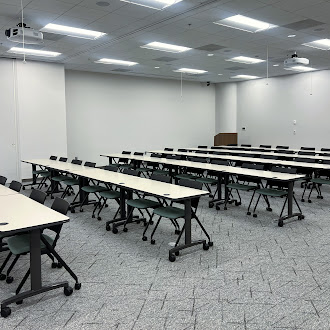 LargeTrainingEventRoom_175Pr_1100_2 Empty modern classroom with rows of wheeled desks and chairs, white walls, and ceiling projectors above.