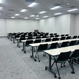 LargeTrainingEventRoom_175Pr_1100_3 Empty classroom or lecture hall with rows of white desks and black chairs arranged in a grid under bright ceiling lights.