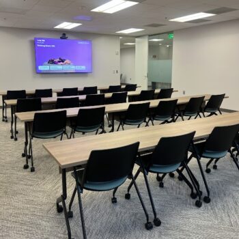Training room with rows of light wood tables and black rolling chairs, facing a purple presentation screen at the front.