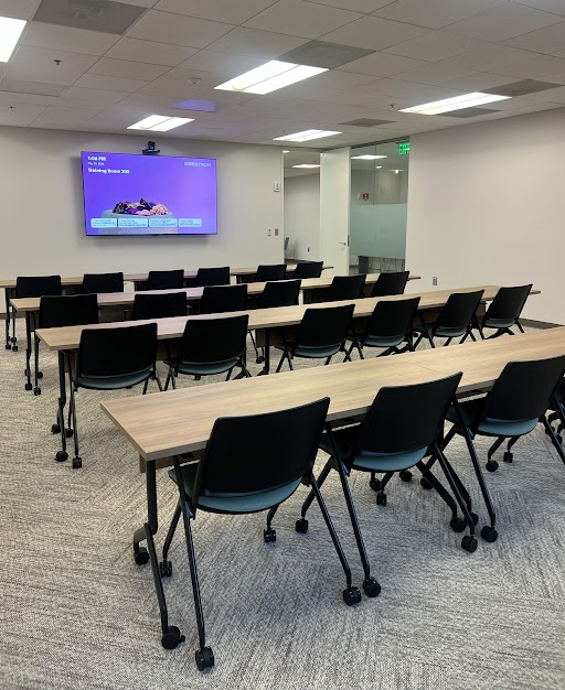 TrainingRoom_24Pr_1100 Training room with rows of light wood tables and black rolling chairs, facing a purple presentation screen at the front.
